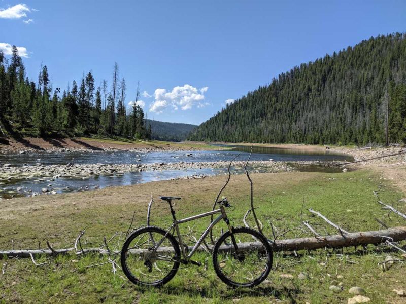 bikerumor pic of the day a bicycle is in a field next to a fallen tree there is a river beyond and a hill with pine trees growing on it the sky is clear and blue except for a few fluffy white clouds low over the mountain ridge.