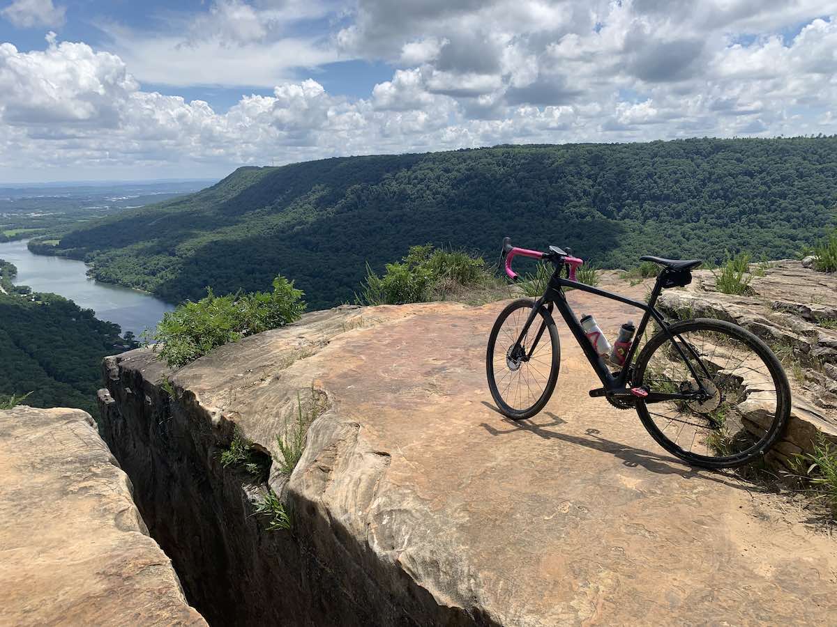 Bikerumor pic of the day a gravel bike is on a large rock slab looking out over green mountains, a river and valley below, the sky is blue and full of white fluffy clouds.