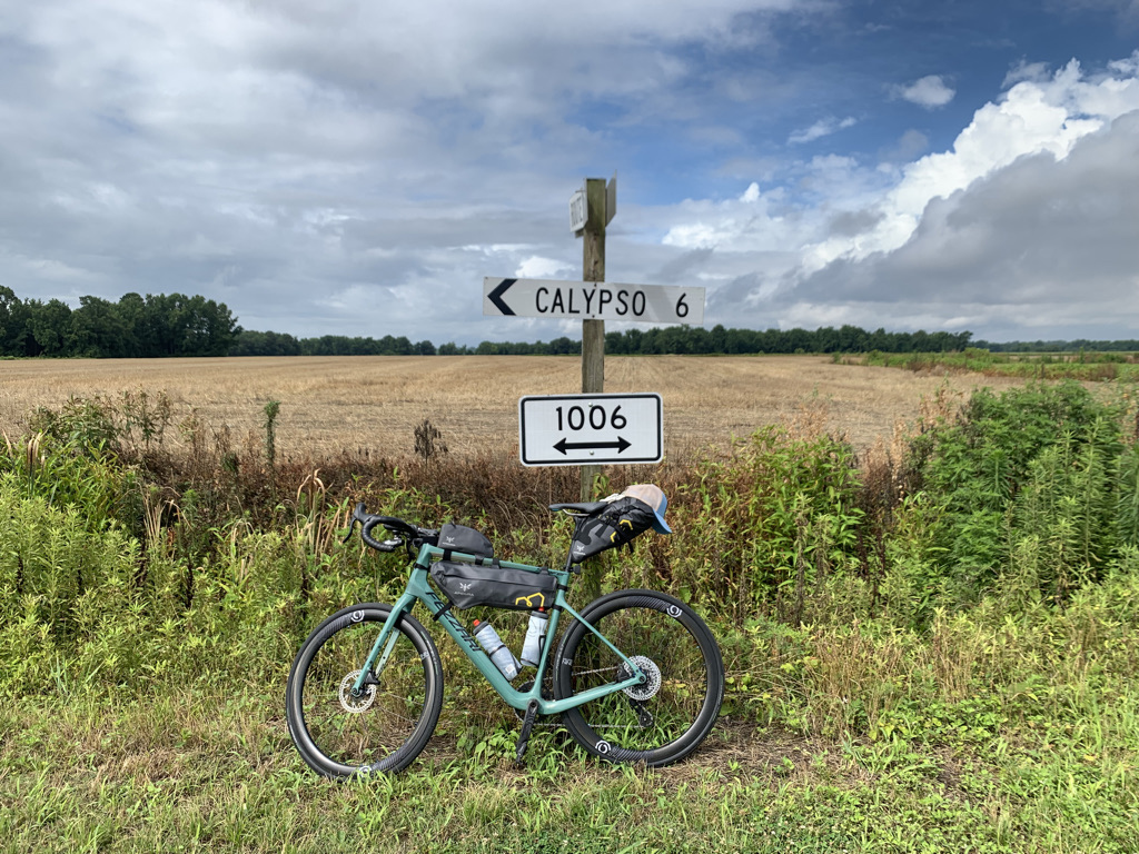 bikerumor pic of the day fezzari gravel bike leaning against a road sign pointing to calypso north Carolina there is a large field of gold behind and a blue sky covered mostly by large fluffy clouds.