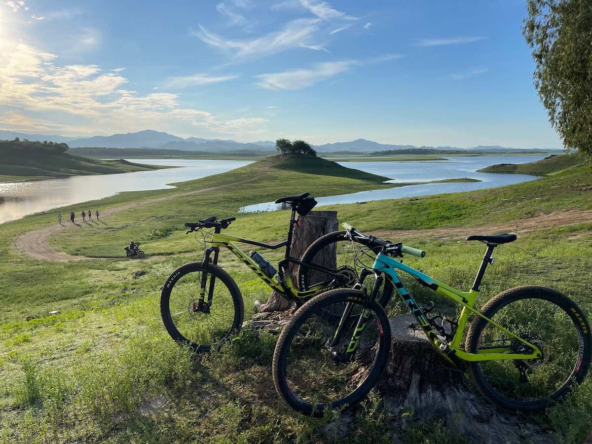 bikerumor pic of the day two mountain bikes lean against a stump in a gently sloping grassy field, there is a lake in the distance and there are whips clouds in the blue sky.