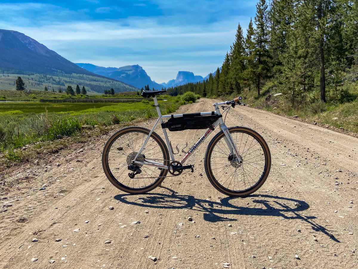bikerumor pic of the day a gravel bike is displayed across a wide gravel and dirt road with a pine forest on the right and a green valley on the left with views of the tetons in the distance. the sky is bright and the sun is directly overhead.