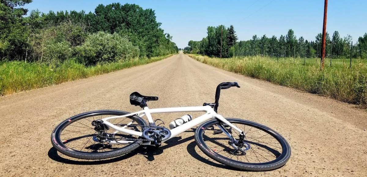 bikerumor pic of the day a bicycle is laid on a gravel road the perspective is low and makes the road look like it goes straight forever and disappears, there is green grass and scrub brush neither side of the road and the sky is clear.