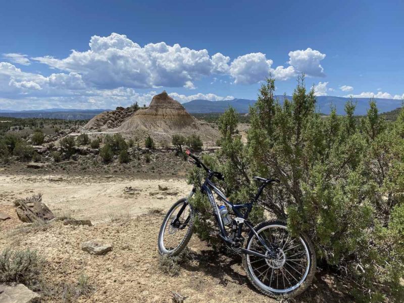 bikerumor pic of the day a bicycle leans against some scrubby bushes near a dirt trail, the sky is blue with fulffy white clouds in the distance and there are eroded rock formations in the background.