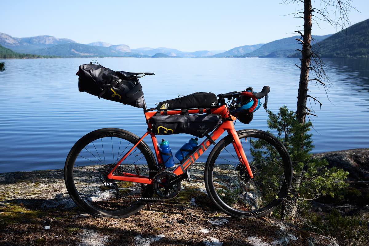 bikerumor pic of the day a bicycle has bike packing bags on it and is posed in front of a large calm lake with mountains surrounding it.