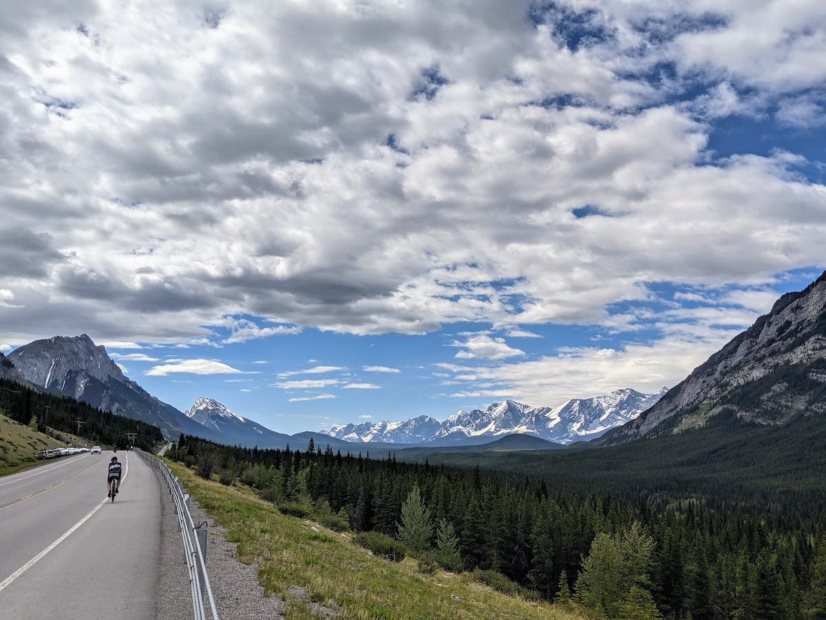 bikerumor pic of the day a cyclist rides on highway 40 towards the highwood pass in alberta canada the rocky mountains of banff are snowcapped and in the distance, the sky is blue with cotton candy clouds