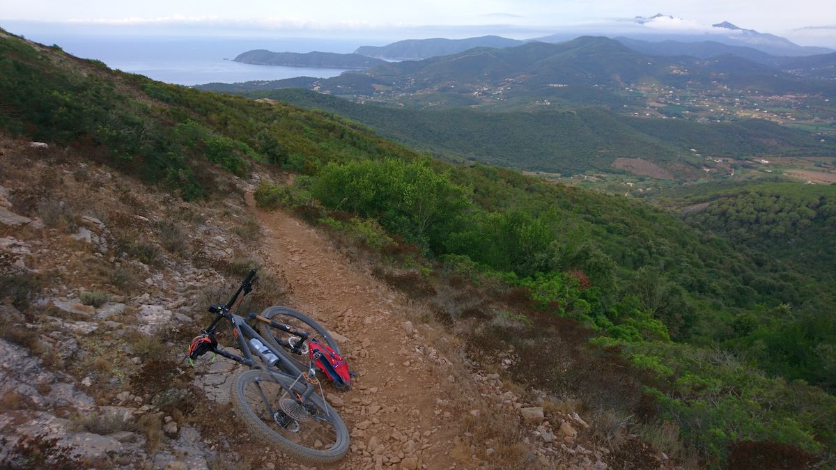 bikerumor pic of the day a mountain bike leans on the side of a dirt trail on the side of a mountain overlooking the rest of the island with a body of water in the distance.