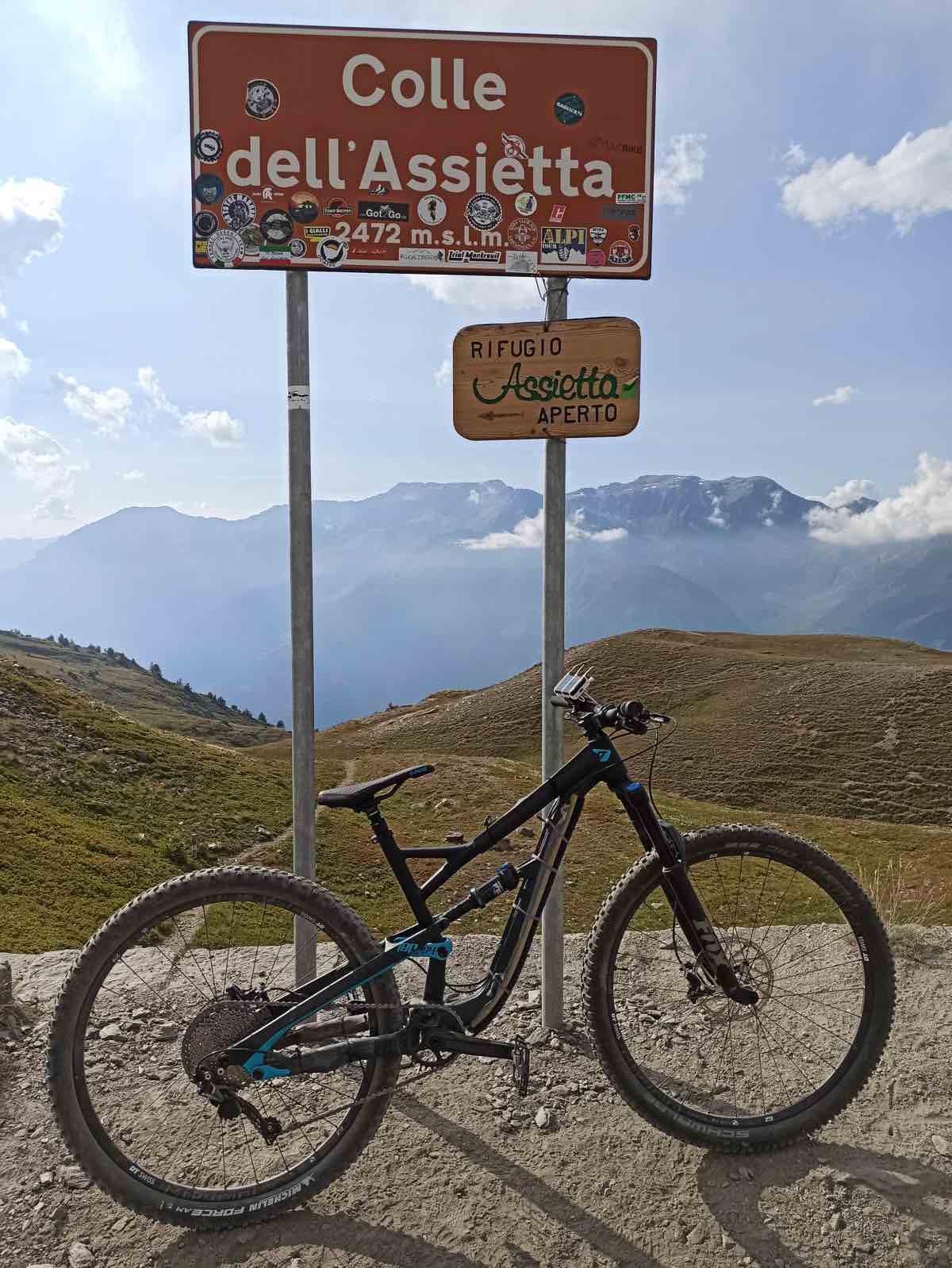 bikerumor pic of the day a mountain bike is next to a sign that says Colle dell'Assietta and a sign for a refugio on the top of a mountain pass in the alps, the mountains in the distance are covered by clouds.