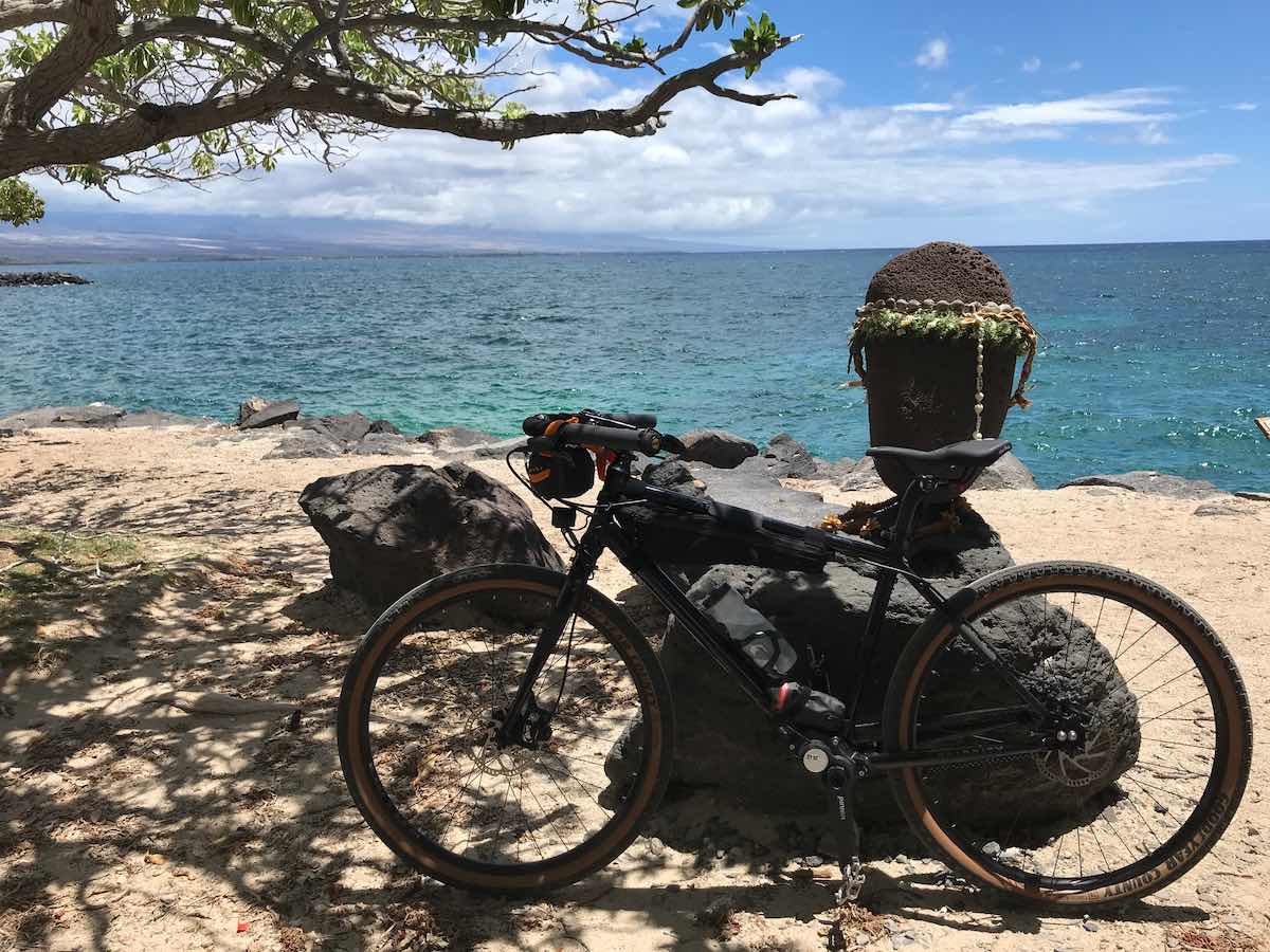 bikerumor pic of the day a bicycle leans against volcanic rock on white sandy beach and blue green ocean waters.