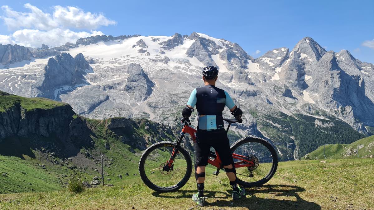 bikerumor pic of the day a cyclist stands with their bike overlooking a glacier that looks like a grey rocky mountain, the cyclist is standing on green grass and the sun is high in the sky
