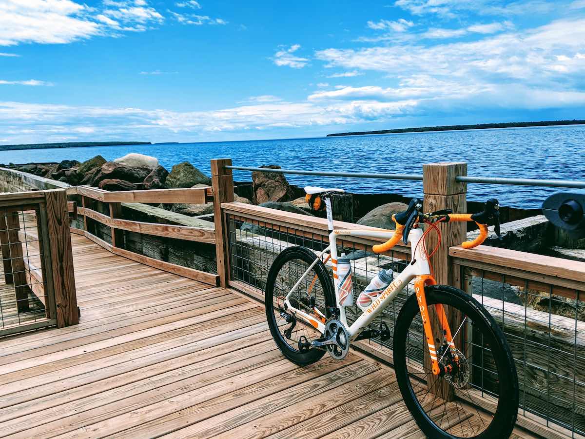 bikerumor pic of the day sand point wisconsin a bicycle leans against the wooden fence of a dock leading out over lake superior with small islands in the distance and partial clouds in the blue sky