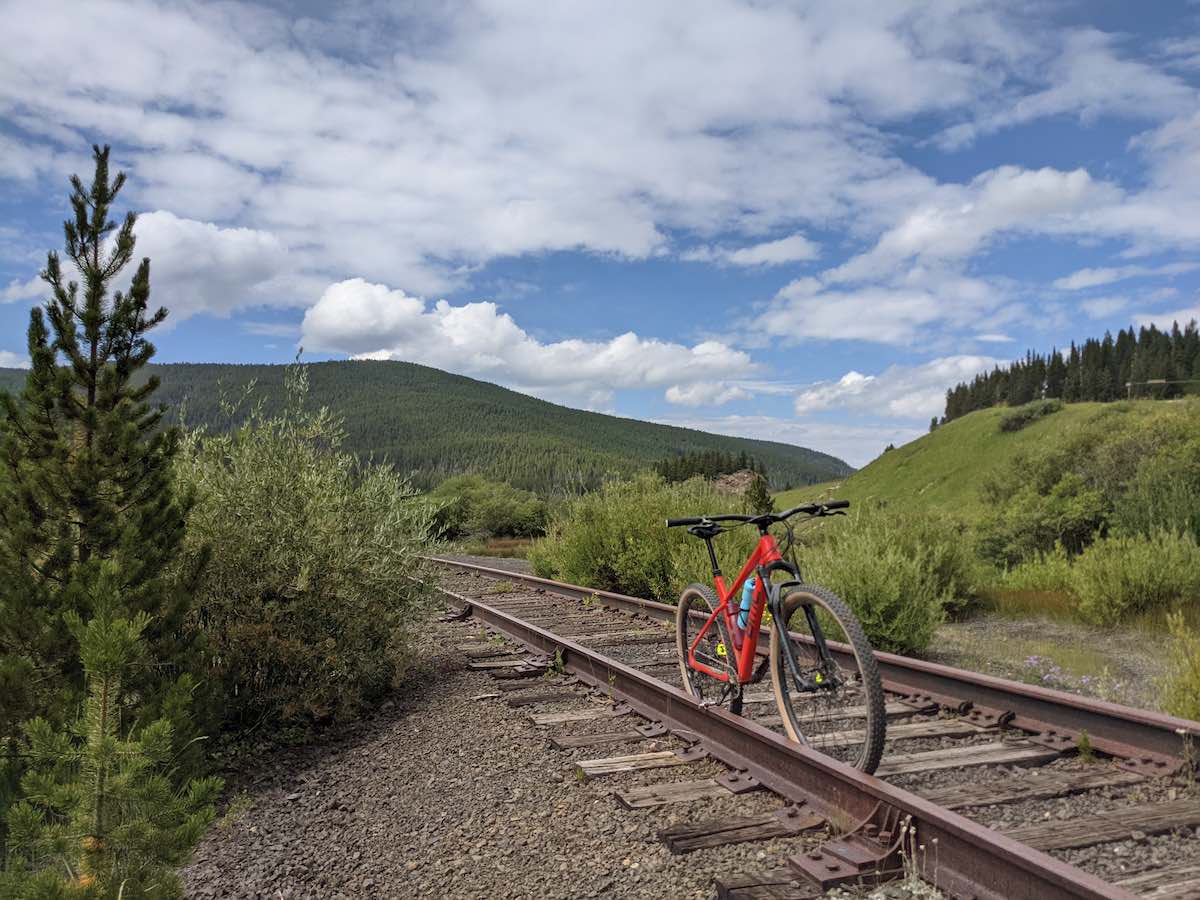bikerumor pic of the day a red mountain bike is on an old trail rail line in colorado with sage brush on either side and a mountain in the distance, the sky is blue with fluffy clouds.
