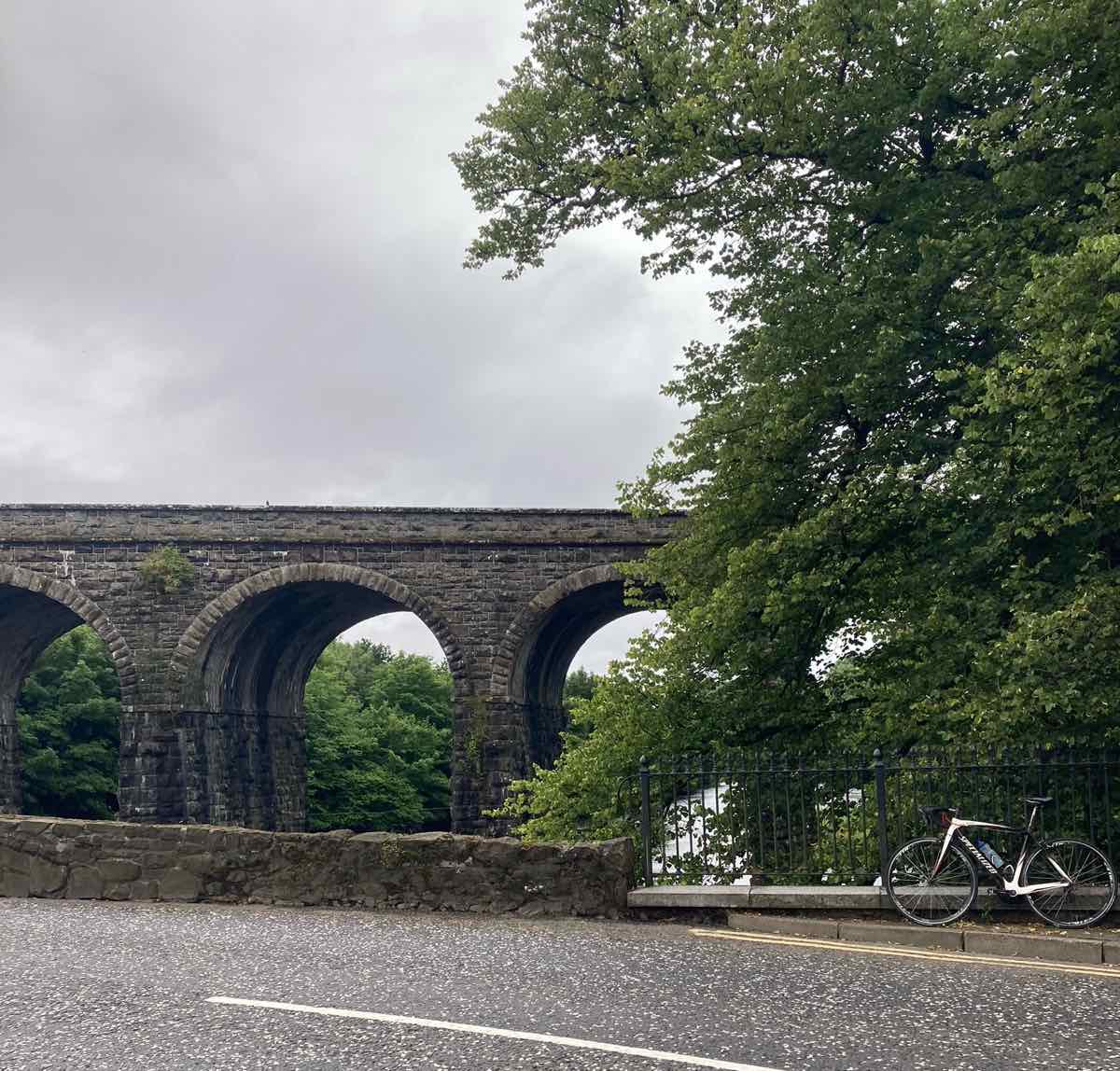 bikerumor pic of the day a road bike leans against a low stone wall the borders the road with a stone arched bridge in the distance. the sky is cloudy and bright.
