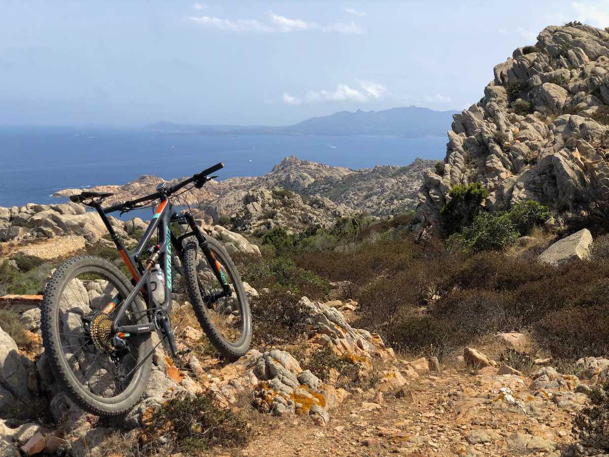 bikerumor pic of the day a mountain bike leans agains a large outcropping of jagged rock on the island of Cabrera off the coast of italy, the ocean is down in the distance and the sun is high overhead.