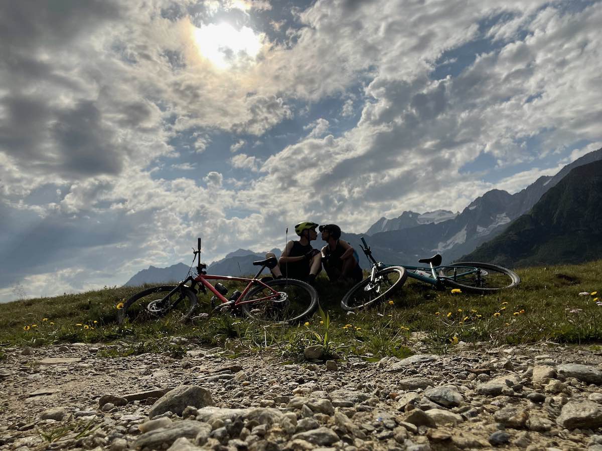 bikerumor pic of the day a couple sits together on the rocky ledge of a mountain with their mountain bikes laid at their feet, the sky is partly cloudy and the sun is strong behind them.