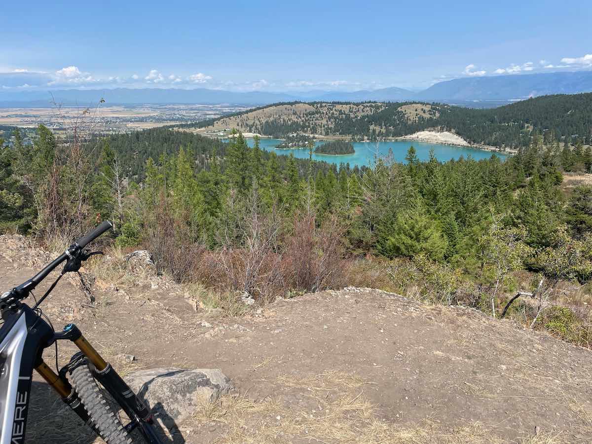 bikerumor pic of the day a bike is seen in the corner of the picture on a dirt landing overlooking a clear blue lake on the side of a mountain surrounded by pine forrest and a small town in the distance, the sky is blue and the sun is high overhead.