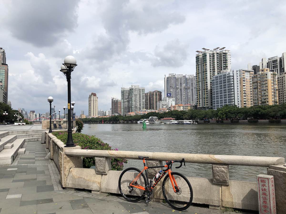 bikerumor pic of the day a red bike leans a against a concrete railing along the pearl river, there are tall buildings across the river and the sky is filled with clouds.