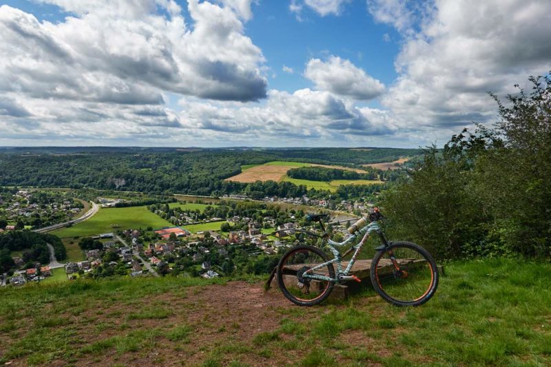 bikerumor pic of the day a mountain bike on a grassy clearing looking out over a verdant valley with a small village, the sky has fluffy white clouds with peeks of blue sky and the sun is high and bright.