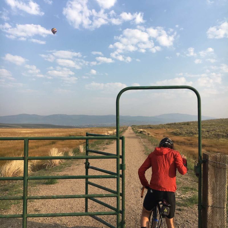 bikerumor pic of the day a cyclist passes through a livestock gate to access a gravel road that runs straight as far as the eye can see, there are a smattering of fluffy clouds in the sky as well as a hot air balloon in the distance.