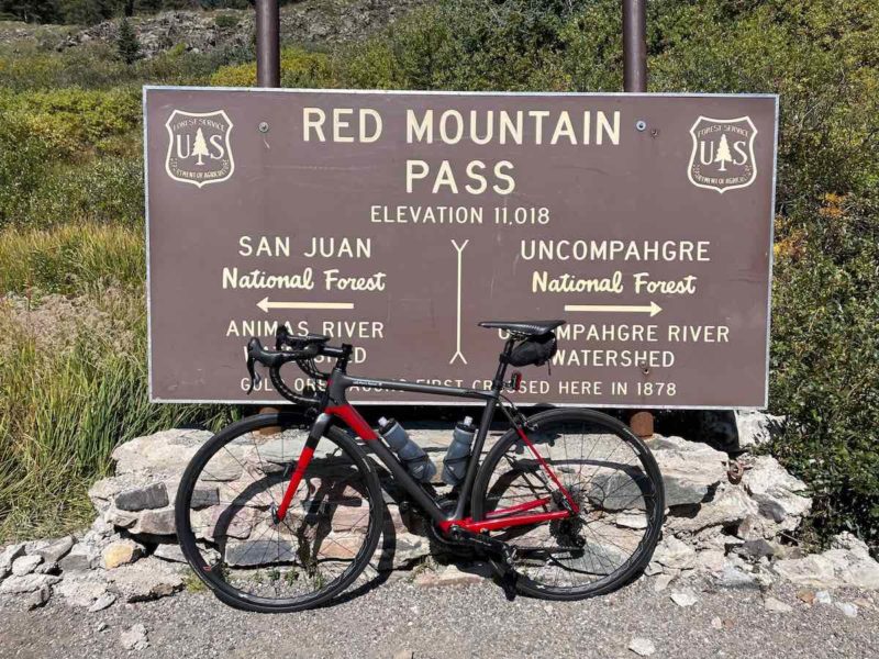 bikerumor pic of the day a red bicycle leans against a sign for the red mountain pass in colorado, the day is sunny and bright and there is green foliage behind the sign.