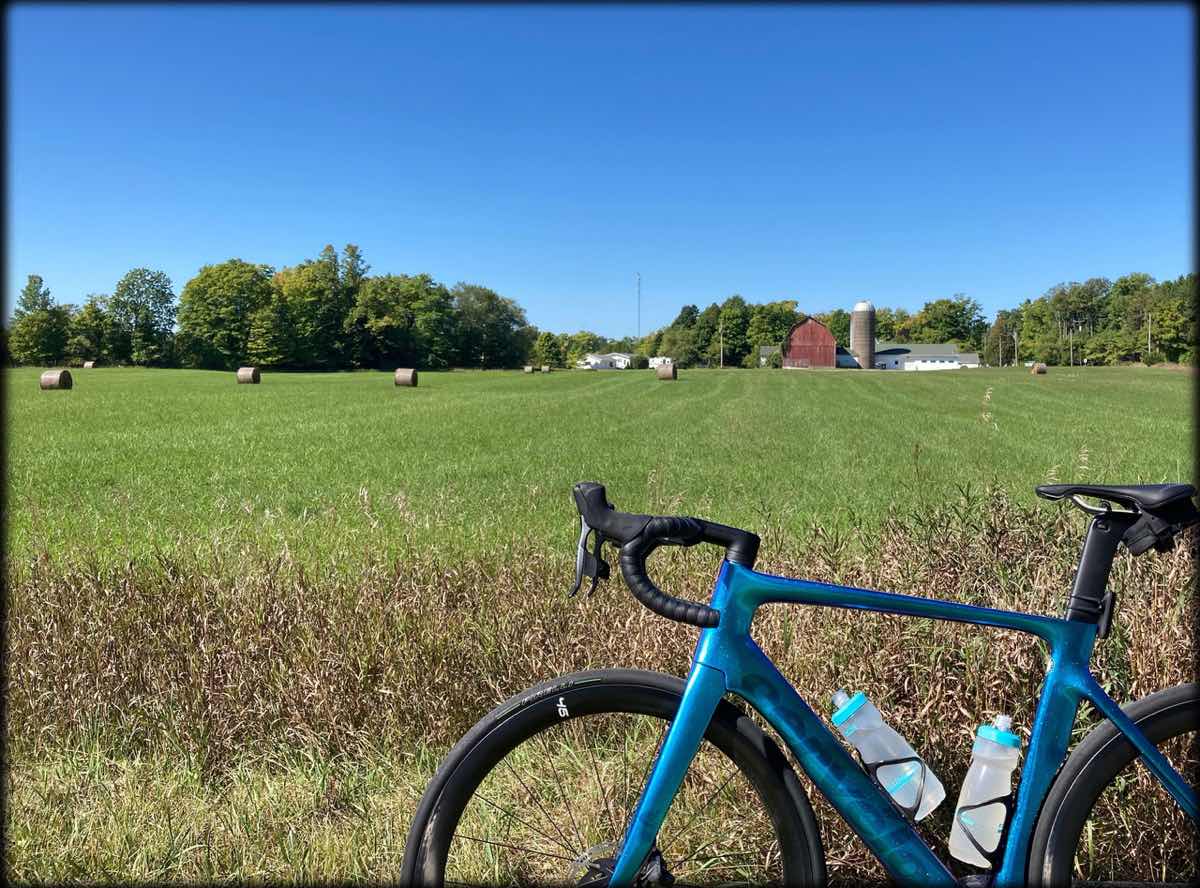 bikerumor pic of the day a blue road bicycle is positioned in front of a green grassy field with bales of hay in the distance and a red barn at the treelike. the sky is clear and blue and the sun is high.