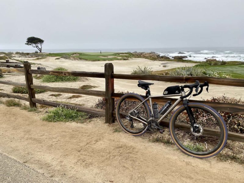 bikerumor pic of the day a bicycle leans against a wooden fence looking out over a sandy dunes and the edge of a golf course. a lone cypress tree is in the distance with the ocean beyond that, the sky is grey and hazy in the early morning light.