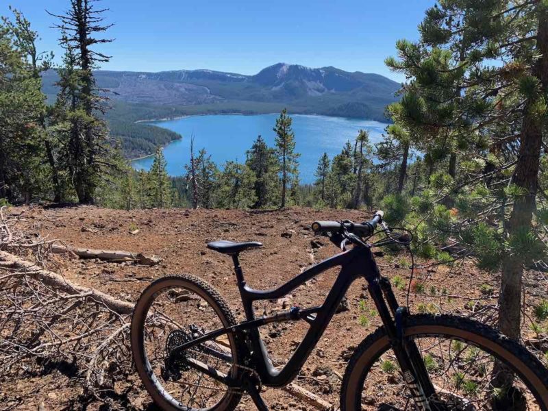 bikerumor pic of the day a mountain bike is positioned near an opening between the pine forest lookin gout onto a crater lake, the sky is clear and the sun is high in the sky.