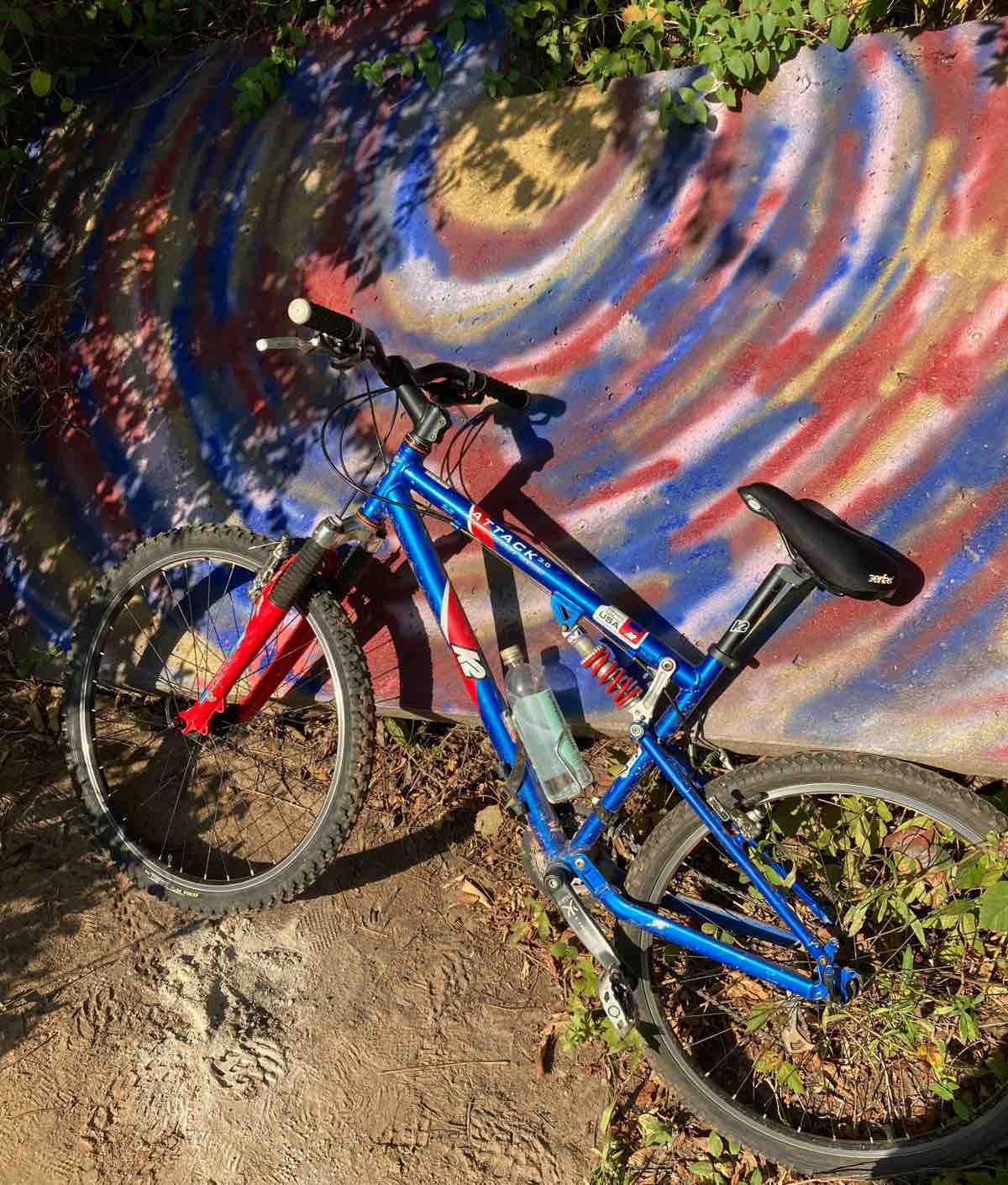 bikerumor pic of the day a blue K2 mountain bike with red fork leans against a concrete wall with a spray painted swirl design with the same colors.