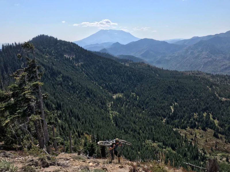 bikerumor Pic of the day a person carrying a mountain bike appears at the top of a trail with pine tree covered mountain peaks in the distance.