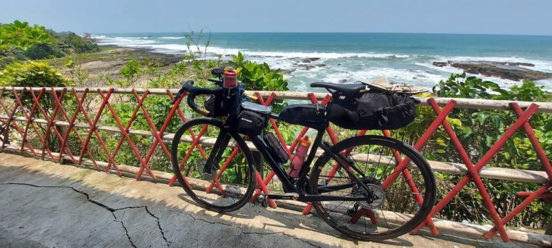 bikerumor pic of the day a gravel bike is packed with gear for a long distance race, it is leaning against a red fence barrier overlooking a rocky beach on the southern edge of indonesia, the sun is bright and the sky is clear.