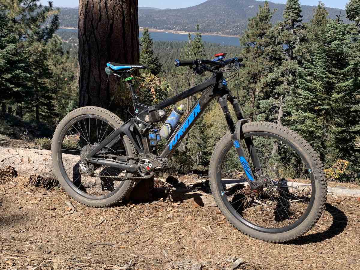 bikerumor pic of the day a mountain bike is on a dit trail next to a large tree trunk, the view beyond the tree are pine trees on the side of the mountain with a view of a lake far away, the sun is bright.