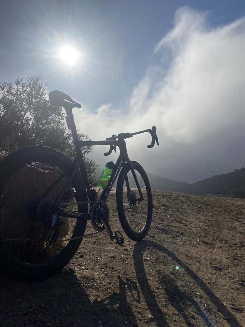 bikerumor pic of the day a bicycle is on a dirt path with a shrub to it's left and a foggy overlook in front of it, the sun is high above and creating a long shadow of the bicycle