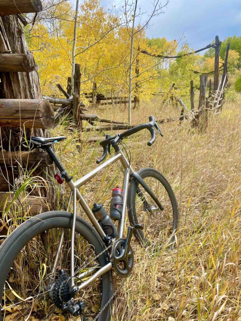 bikerumor pic of the day a bicycle leans against an old wooden structure in a field of golden colored grass, there is a grove of aspen trees in the distance and the leaves are all yellow, there is a tiny bit of sky showing and it is dark with clouds even though it is a bright day.