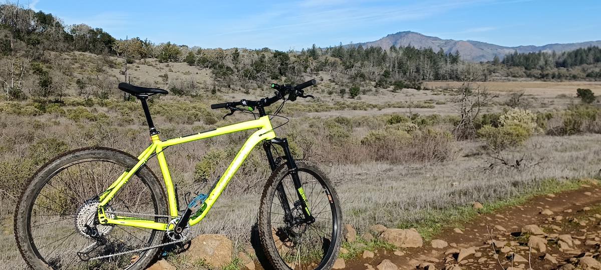 bikerumor pic of the day, a yellow mountain bike is on a dirt trail with a scrub covered hill in the distance, the sky is blue and clear