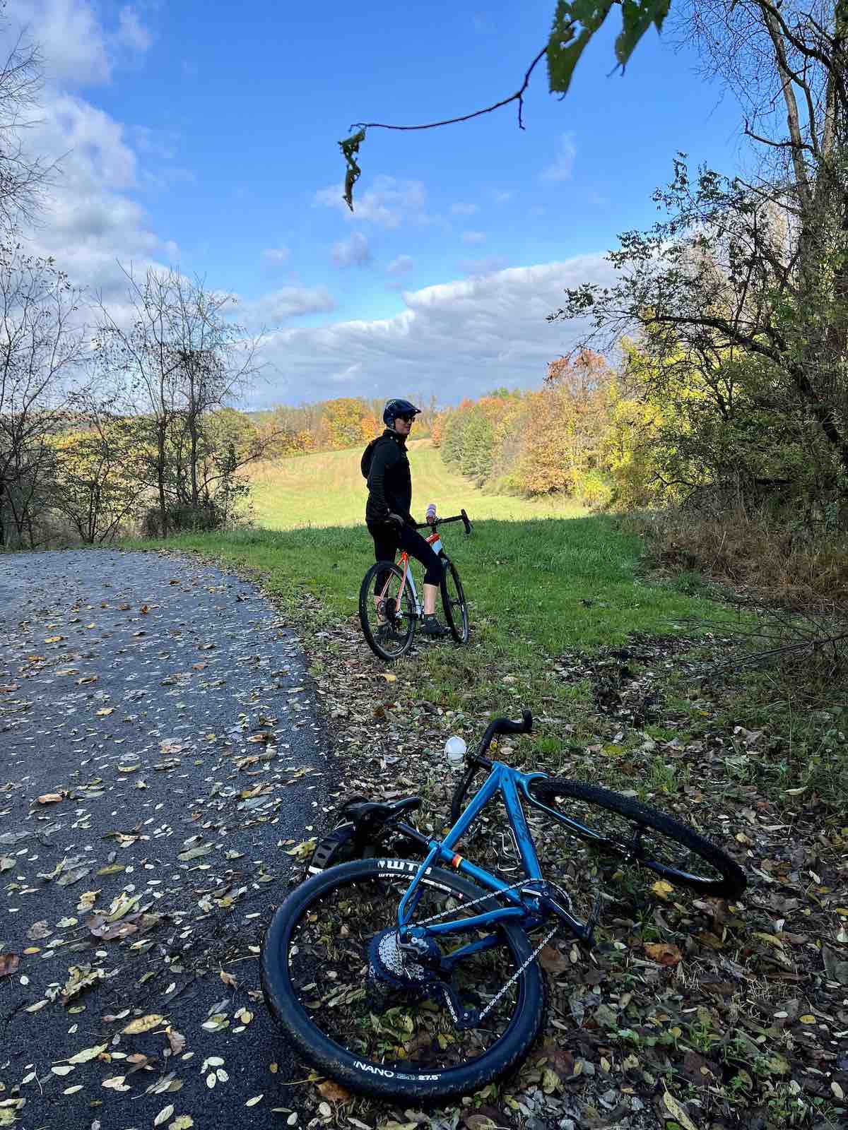 bikerumor pic of the day a cyclist looks back at the photographer whose bike is laying on the ground in front of them beside an asphalt road covered in fall leaves, the grass is green, and the trees beside the road are changing red and yellow, the sky is bright blue with a hint of clouds in the distance.