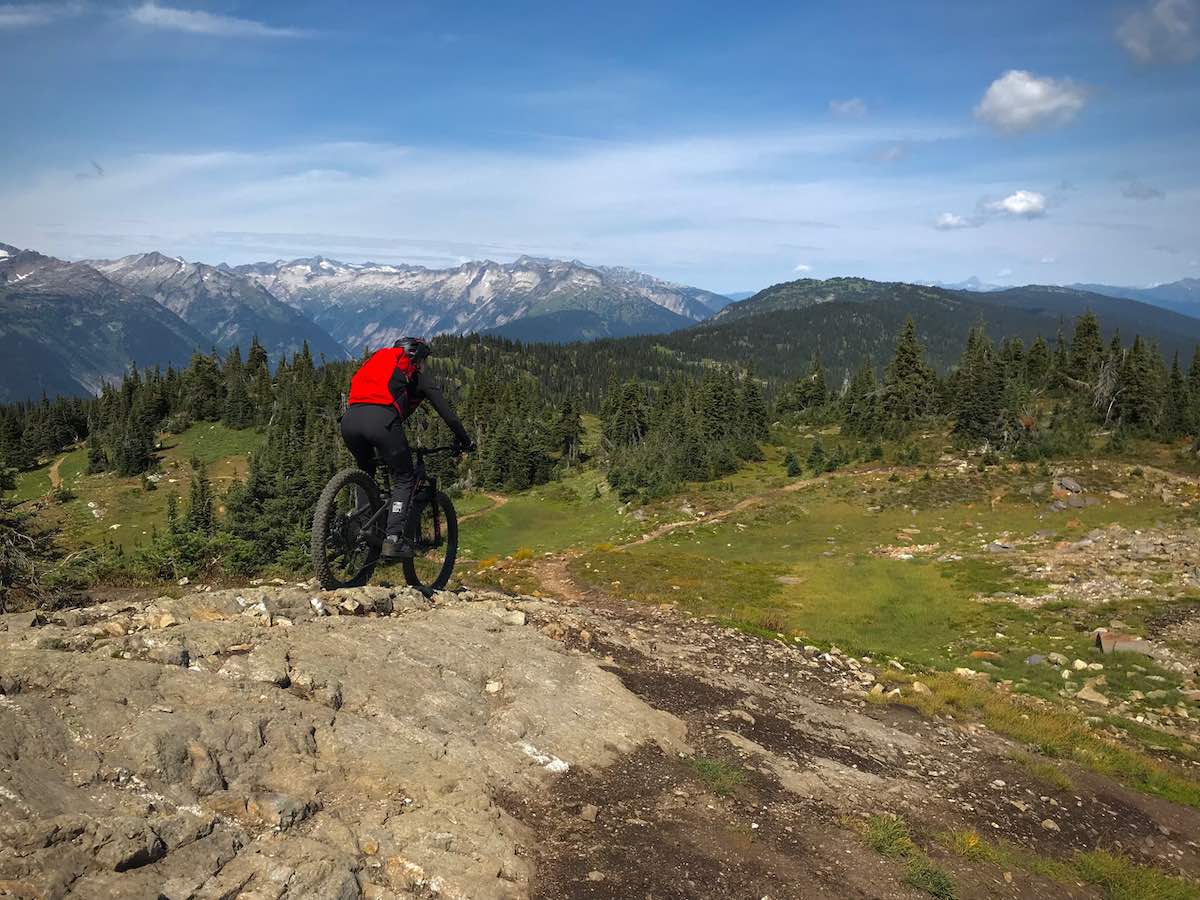 bikerumor pic of the day a mountain biker is on a rocky ridge heading down the slope toward a grassy clearing, the mountains surrounding has pine tree groves and the peak in the distance is covered in snow.