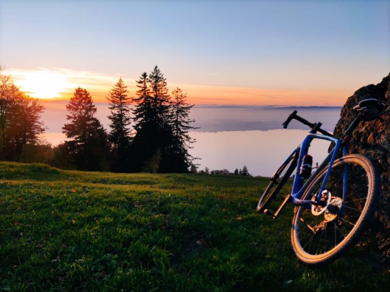 Bikerumor pic of the day a bicycle leans against a rocky outcropping on the top of a mountain, it is green and grassy at the top and overlooks pine trees and clouds covering the land below, the sun is low in the sky and creating a lovely orange glow.