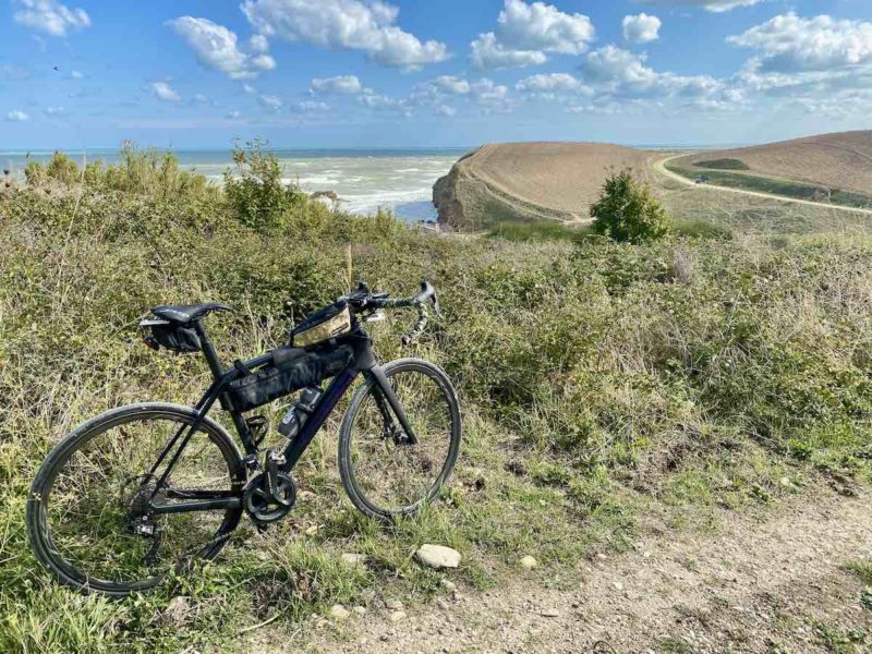 bikerumor pic of the day a bicycle is in some brush along a dirt path looking out over an ocean view with a road and bright blue skies with cotton clouds in the distance.