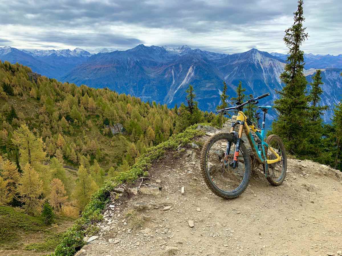 bikerumor pic of the day mountain biking in the alps, a mountain bike is on a steep dirt trail overlooking pine tree forest with blue colored mountains in the distance. The sky is cloudy.