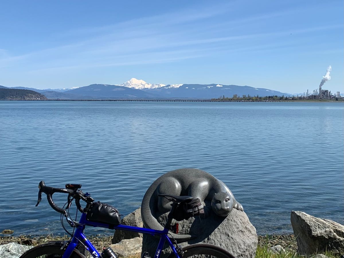 bikerumor pic of the day a bicycle leans against a rock that has a carving of an animal on it, beyond is a large body of water with a snow capped mountain on the other side in the distance, the sky is blue with small whisky clouds.