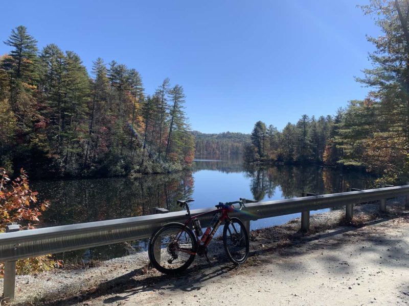 bikerumor pic of the day a bicycle leans against a metal railing along a gravel road looking out over a small lake bordered by pine trees and trees with yellow and orange leaves, the sky is clear and blue.