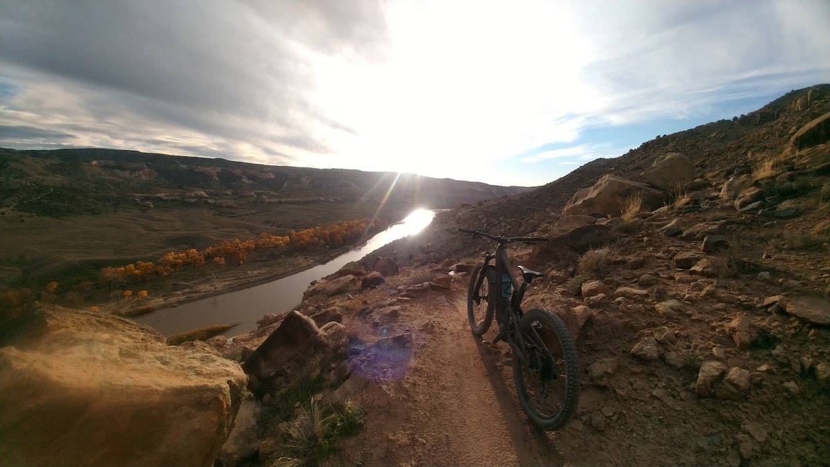 bikerumor pic of the day a mountain bike leans against a rock on a dirt trail that is heading toward the horizon around the side of a hill, the sun is setting on the horizon creating bright rays that almost blind the viewer, the land surrounding the trail is brown dirt and there is a small river hugging the hill below the trail.