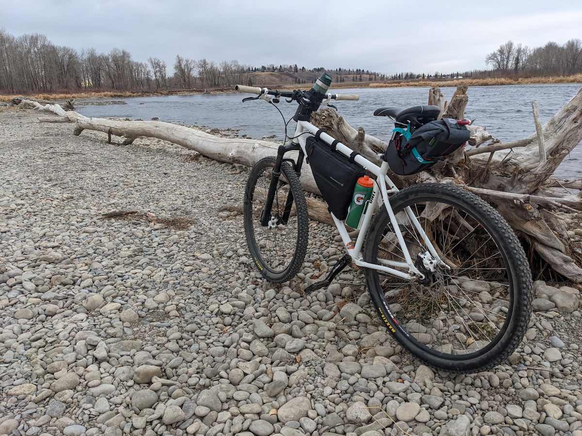 bikerumor pic of the day a bicycle leans against a weathered tree and root system that has washed up on the rocky shore of a lake, the sky is bright but covered in clouds, the land alongside the lake is covered in bare trees.