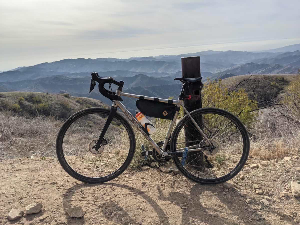 bikerumor pic of the day a lynsey bicycle leans against a marker post on the top of a rocky mountain pass with scrub brush on the edge the trail. the sky is covered in smooth clouds and the sun is bright behind them.