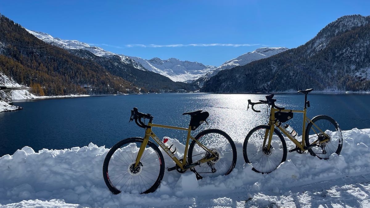 bikerumor pic of the day two gold santa cruz gravel bikes are in the snow beside a large lake with mountains surrounding, the sky is blue and clear and the sun is low in the sky reflecting in the lake.
