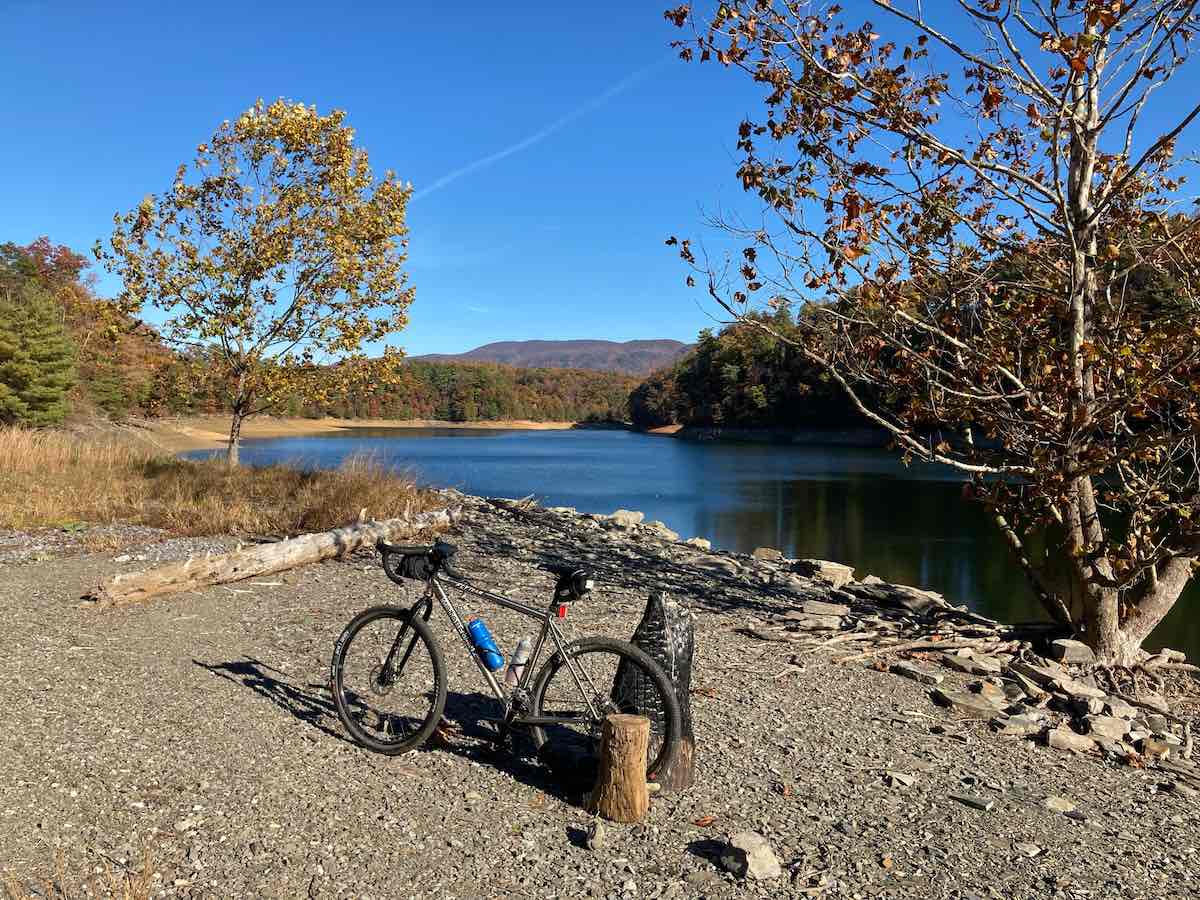 bikerumor pic of the day a bicycle leans against a piece of wood on a gravel landing by a lake, there are trees surrounding the lake and the sun is low in the sky, the leaves are turning brown and gold for autumn and the sky is clear and dark blue.