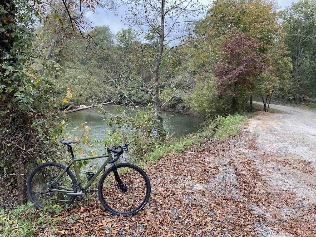 bikerumor pic of the day a bicycle is positioned on the side of a gravel road among dried fallen leaves, there is a small pond alongside the road and trees surrounding them that are changing their colors for autumn.