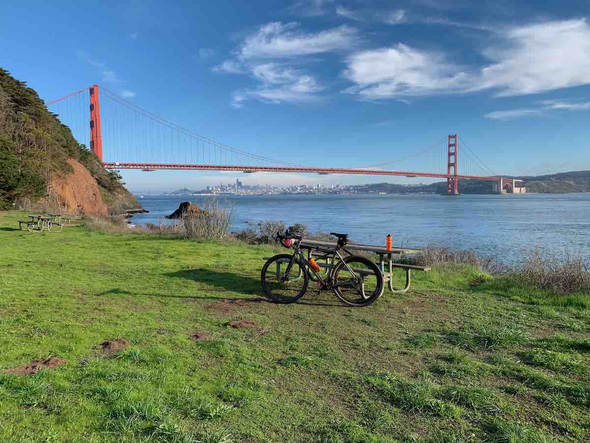 bikerumor pic of the day a bicycle leans against a picnic table on a grassy clearing overlooking the golden gate bridge, the sky is bright and blue and there are a few wispy clouds in the sky.