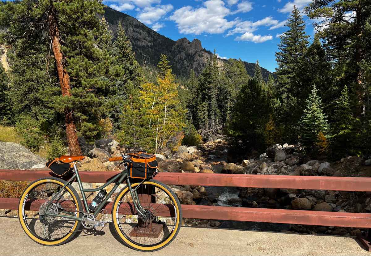 bikerumor pic of the day a touring bicycle is on a wooden bridge overlooking a large stream with boulders surrounding it, pine trees along the stream and up the mountain and rocky ridges beyond, the sky is blue with some fluffy white clouds, the day is sunny and bright.