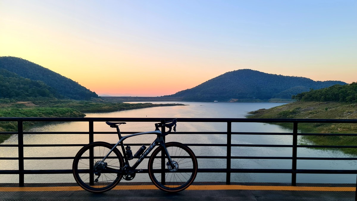 bikerumor pic of the day a bicycle leans against the metal railings of a bridge that spans a body of water surrounded by low mountians, the sun has just disappeared and left an orange glow in the grey sky that reflects in the water.