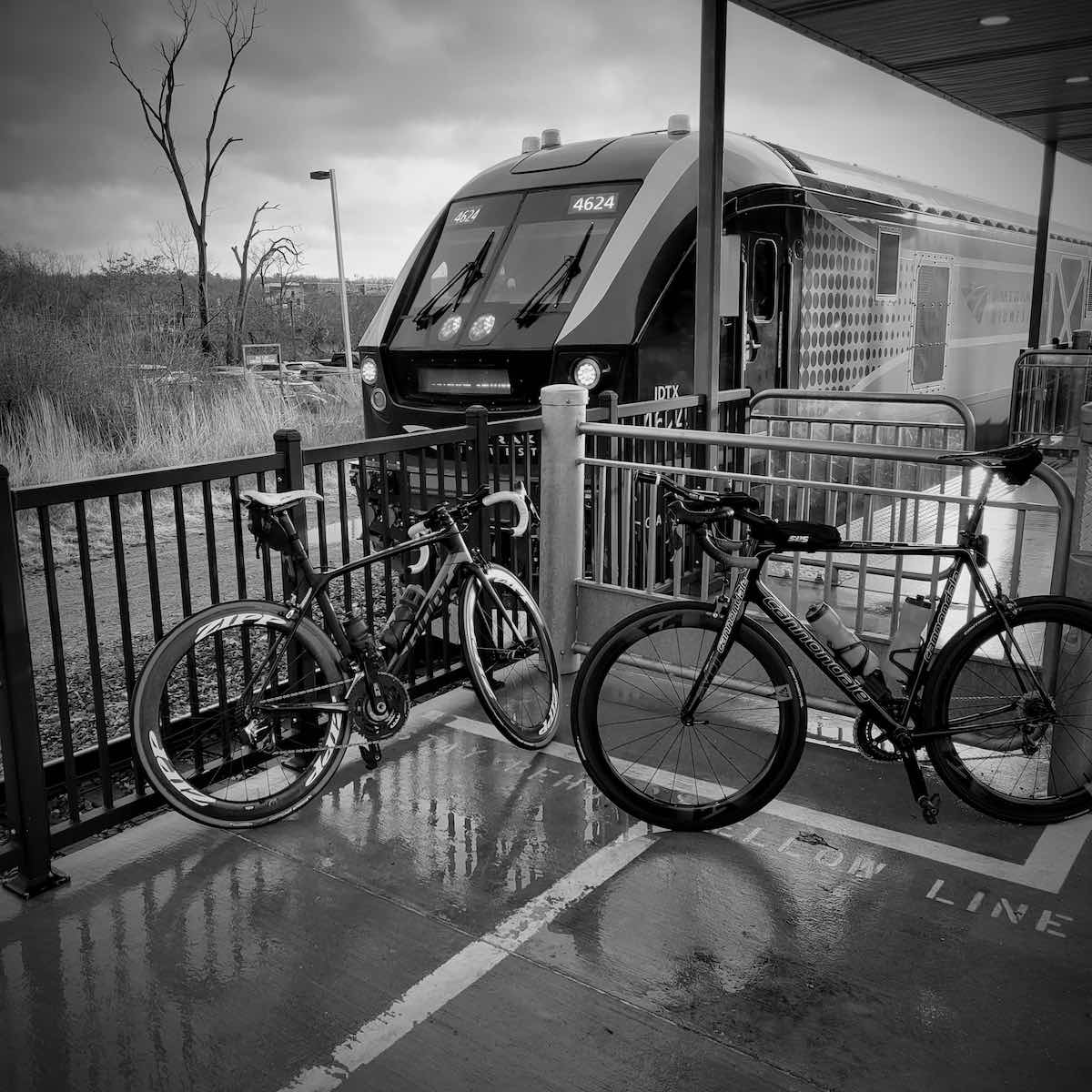 bikerumor pic of the day two bicycles lean against a metal railing on a train platform with an amtrak train behind them, the photo is in black and white and the ground is wet from rain.